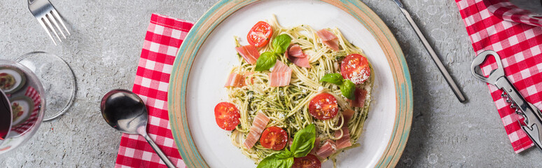 top view of Pappardelle with tomatoes, basil and prosciutto on plate on plaid napkin with cutlery near red wine on grey surface, panoramic shot