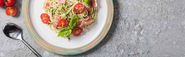 top view of cooked Pappardelle with tomatoes, basil and prosciutto near spoon and tomatoes on grey surface, panoramic shot