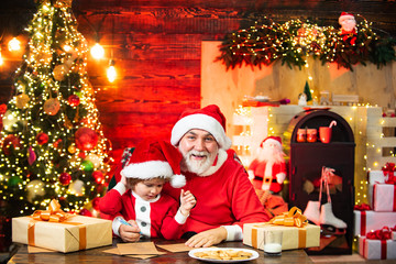 Funny Santa helper boy in Santa hat writes letter with Santa near christmas tree.