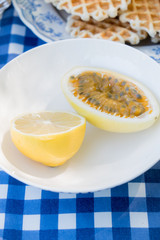 Picnic table decoration on nature with a white plate with lemon and passion fruit and waffles, everything stands on a blue checkered tablecloth.