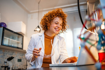Young female scientist working on the computer in a laboratory. Female researcher taking notes at her workplace. She is standing in her laboratory, using microscope and laptop for her research. 