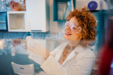 Young attractive female scientist in protective glasses and gloves dropping a liquid substance into the test tube with a long glass pipette in the scientific chemical laboratory