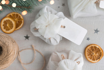 Fototapeta na wymiar Christmas presents wrapped with white furoshiki fabric, labels and dried orange slices. Eco friendly gift.