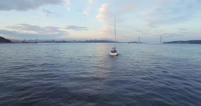 Aerial panoramic view of bautiful white sailing yacht with high mast in the Eastern Bosphorus strait at summer sunset. Vladivostok cityscape and Russian bridge are on the background