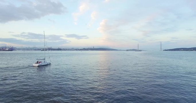 Aerial view of bautiful white sailing yacht with high mast in the Eastern Bosphorus strait at summer sunset. Vladivostok cityscape and Russian bridge are on the background