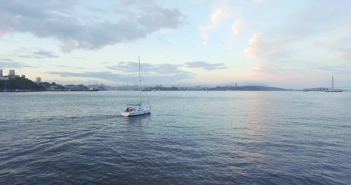 Aerial panoramic view of bautiful white sailing yacht in the Eastern Bosphorus strait at summer sundown. Vladivostok cityscape and Russian bridge are on the background