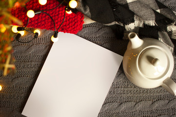 Christmas mockup, with a postcard, wishes, letter on the blanket with a kettle, leaf, tea, cone, chocolate and xmas tree lights. Background top view blank space for text.