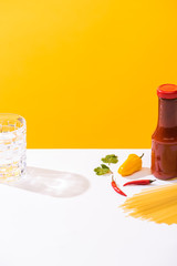Fototapeta na wymiar Bottle of ketchup beside peppers, raw spaghetti and glass of water on white surface on yellow background