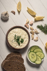 Top view of homemade tzatziki sauce with bread and ingredients on white wooden background