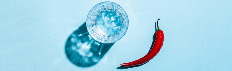 Top view of ripe chili pepper and glass of water with shadow on blue background, panoramic shot