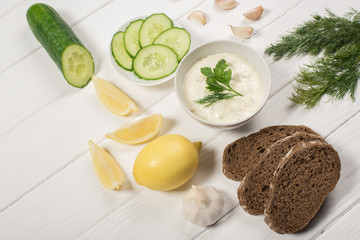 Tasty tzatziki sauce with fresh ingredients and bread on white wooden background