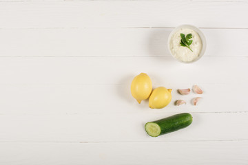 Top view of homemade tzatziki sauce with lemons, cucumber and garlic on white wooden background