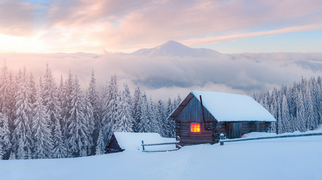 Fantastic winter landscape with wooden house in snowy mountains. Hight mountain peaks in foggy sunset sky. Christmas and winter vacations holiday concept