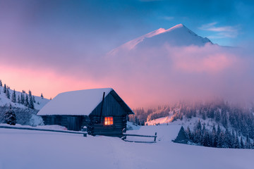 Fantastic winter landscape with wooden house in snowy mountains. Hight mountain peaks in foggy...