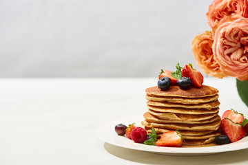 delicious pancakes with blueberries and strawberries on plate near rose flowers on white surface...