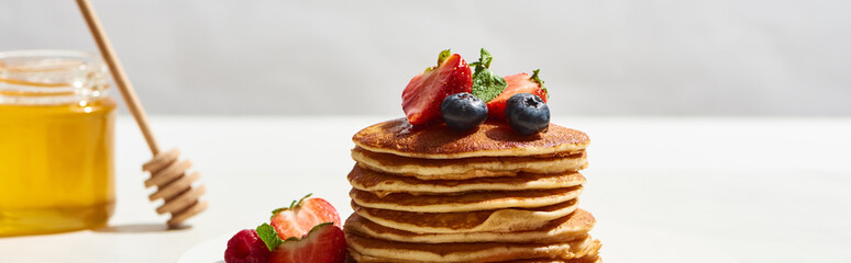 selective focus of delicious pancakes with blueberries and strawberries near honey on white surface isolated on grey, panoramic shot