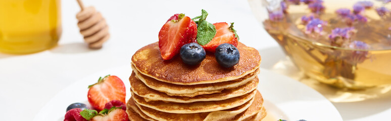 selective focus of delicious pancakes with honey, blueberries and strawberries on plate on white surface, panoramic shot