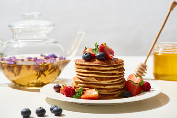 selective focus of delicious pancakes with honey, blueberries and strawberries on plate near herbal tea on white surface isolated on grey