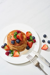 top view of delicious pancakes with maple syrup, blueberries and strawberries on plate near fork and knife on marble white surface
