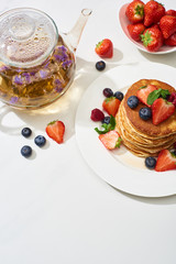 top view of delicious pancakes with maple syrup, blueberries and strawberries on plate near herbal tea in teapot on marble white surface