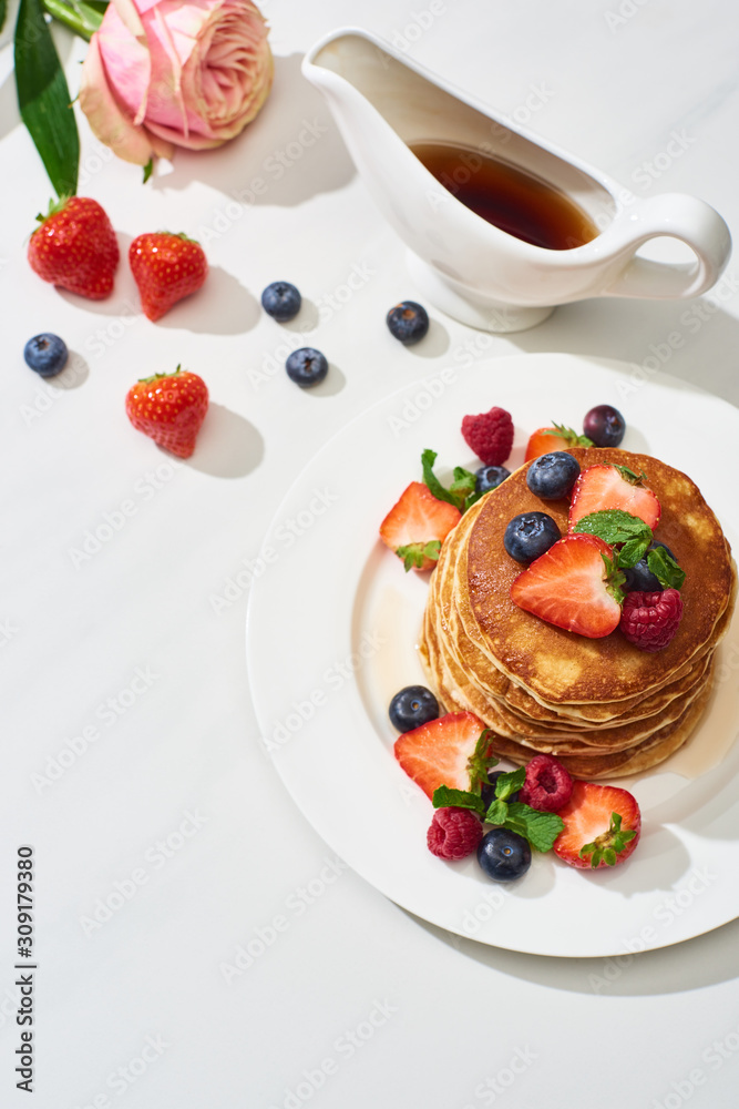 Wall mural top view of delicious pancakes with maple syrup, blueberries and strawberries on plate near rose flower on marble white surface