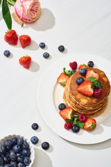 top view of delicious pancakes with blueberries and strawberries on plate near rose flower on marble white surface
