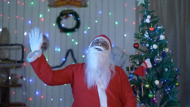 Old funny Santa laughing out loud in glass spectacles on Christmas Eve in India. Happy Santa Claus waving his hand while making a ho ho ho sound during Christmas celebration near a decorated Christ...