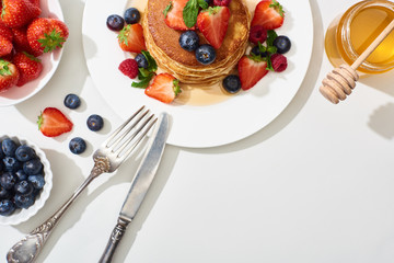 top view of delicious pancakes with honey, blueberries and strawberries on plate with cutlery on marble white surface