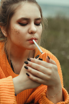 A young woman in a yellow sweater smokes a cigarette with collected hair and looks into the distance. Sits in a green meadow.