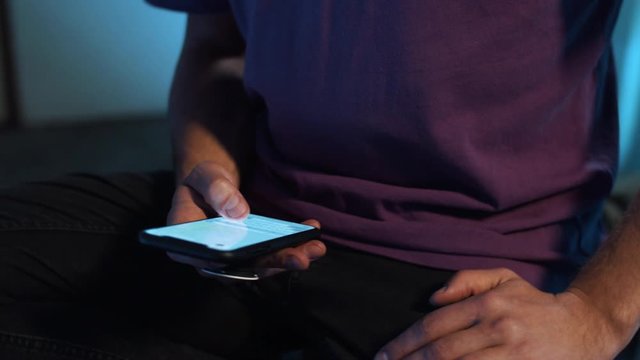 Mature guy in violet t-shirt with bigh watch is sitting in room with dimmed light and holding smartphone in hands, actively using it , while camera is automatically moving around him. Rotation video.