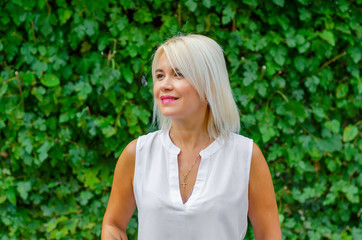 Beautiful blonde middle-aged woman in green leaves background. In white blouse, standing on street. Pretty smile on face straight short hair, brown eyes