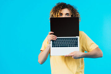 scared curly student hiding behind laptop with blank screen isolated on blue