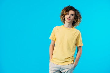 curly teenager with hands in pockets isolated on blue