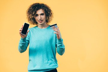 curly teenager holding smartphone and credit card isolated on yellow