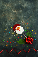 Christmas or New Year background, top view of a Santa Claus lollipop, two branches of thuja or spruce, red bow and multi-colored star-shaped confetti on a dark concrete surface. Empty space for text