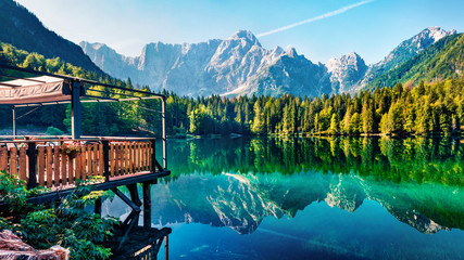 Cozy cafe on the pier. Calm morning view of Fusine lake. Colorful summer scene of Julian Alps with Mangart peak on background, Province of Udine, Italy, Europe. Beauty of nature concept background.