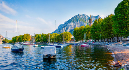 Fototapeta na wymiar Blue waters of the Alpine lake are sparked in the first rays of the sun. Amazing summer scene of Como lake. Wonderful morning cityscape of central park of Lecco town, Italy, Europe.