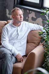 Fototapeta premium Pensive caucasian successful middle-aged man sits on a sofa in a comfortable room and enjoys the weekend. Positive male businessman thinks about future prospects