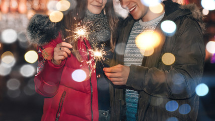 Young smiling couple is having fun with sparklers in winter city nigh