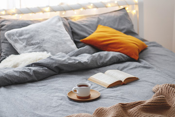 Cup of tea and book lie on the bed with gray linen, blanket, pillows, plaid and led garland light....