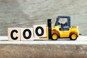 Toy forklift hold letter block O to complete word COO (abbreviation Chief operating officer) on wood background