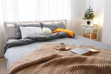 Christmas cozy winter home scene. New year interior decorations. Stylish cosy bedroom decor. Christmas tree on bedside table and bed with grey linen, blanket, pillows, knitted plaid, cup of tea, book.