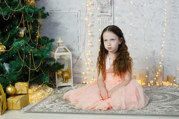 Little cute sad girl sitting at the Christmas tree. New Year room with at home. Christmas good mood. Lifestyle, family and holiday.