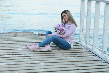 A woman with flowers in her hands sits on a wooden pier near the water on the background of the mountain.