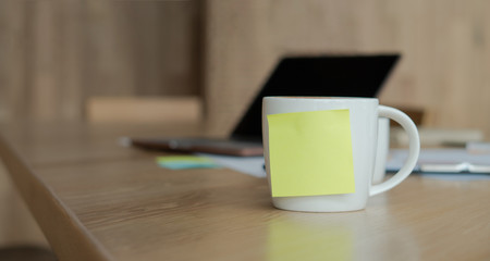 A white coffee cup with a blank yellow Sticky Note on the cup. Resting on the desk and laptop is placed on the back.