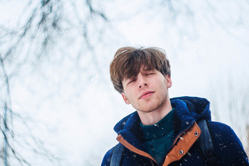 Portrait of a handsome young man who enjoys a winter walk and snowfall in the forest. Walking in the snowfall guy with eyes closed with pleasure.