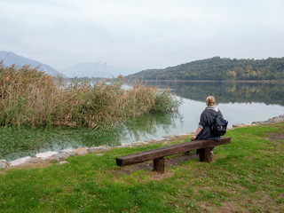 Fototapeta na wymiar Lonely woman sitting on the bench by the lake in the middle of the mountains. Gray autumn day bench