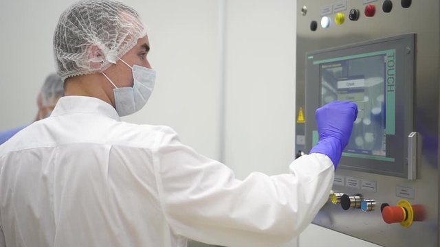 Man operator watch and press on screen during working day in pharmaceutical factory. Male worker works with industrial equipment in pharma chemical company. Employee is wearing white uniform. Concept