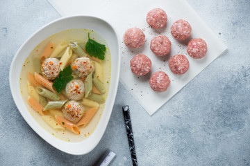 Soup with meatballs and penne pasta, flatlay over light-blue stone background with raw meatballs