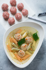 Meatball soup with penne pasta served in a grey bowl, vertical shot on a light-blue stone surface with raw meatballs in the background